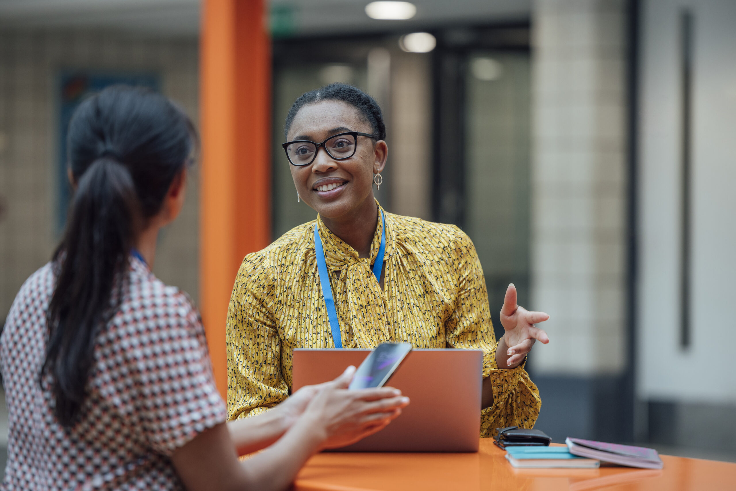 Two female teachers sitting at a table, having a discussion about change management in schools in the lobby area in the school they work at near New York. They are talking about school leadership development together while using a laptop.