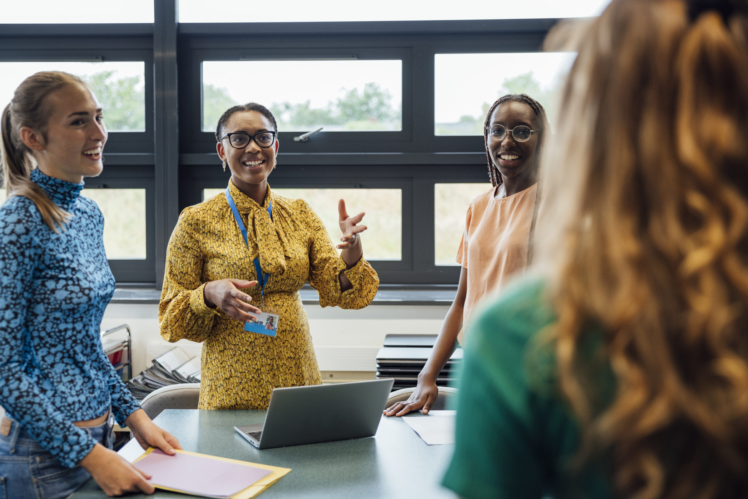 A shot of a mid-adult secondary school teacher talking in a group. They are wearing casual clothing and discussing what they learned in leadership coaching for educators, in a school in New York.