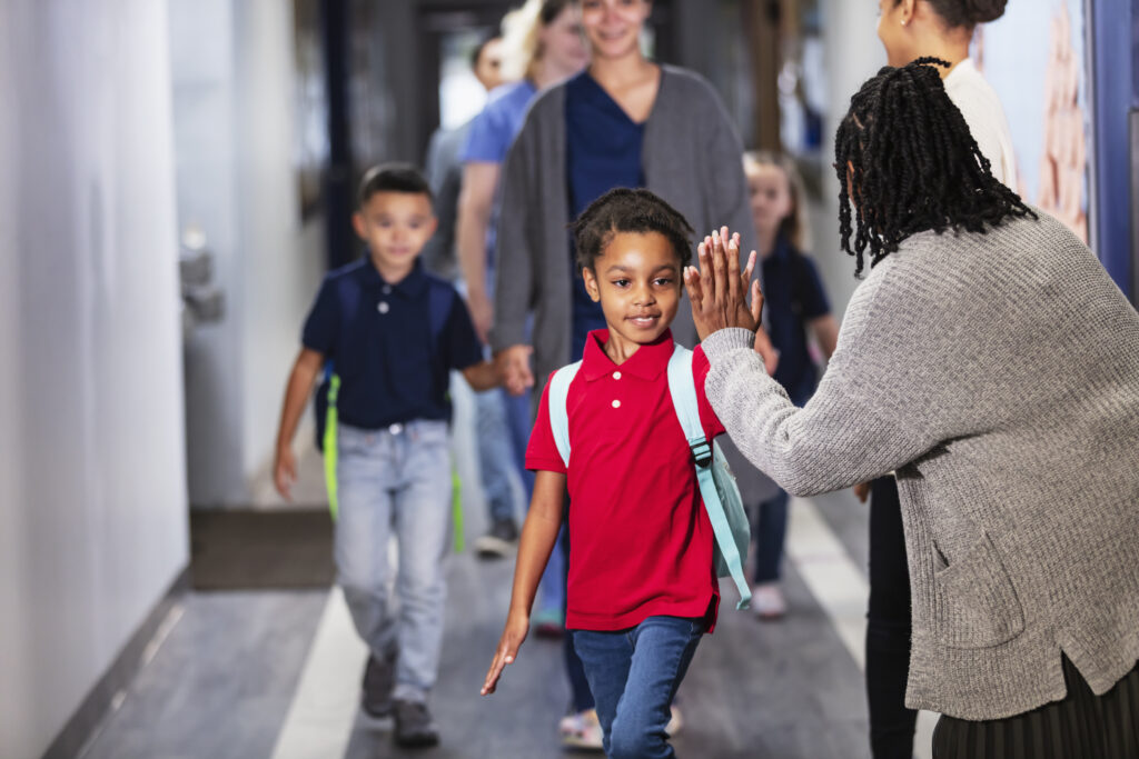 A multiracial group of parents dropping their children off inside an elementary school. The teacher, a black woman in her 30s, is giving a 7 year old black and Hispanic girl a high-five. The main focus is on the girl and the teacher, and the view is from over teacher's shoulder. This is the hero image for Change-Management Resources & Leadership Tools for Educators, focusing on managing change in schools.